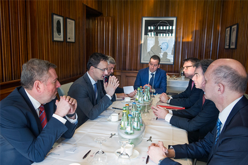 Seven men in suits sit at a decked table